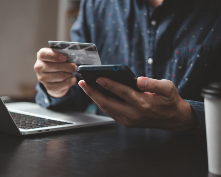 En la imagen, se puede ver a una persona realizando una compra en línea. La persona sostiene una tarjeta de crédito en una mano y un teléfono móvil en la otra, mientras que una computadora portátil está abierta en la mesa frente a ellos. La escena sugiere un entorno de trabajo o estudio, con una taza de café visible en el fondo, creando un ambiente cómodo y productivo. Este momento captura la conveniencia y facilidad de las transacciones electrónicas en la era digital, destacando la integración de la tecnología en las actividades cotidianas.